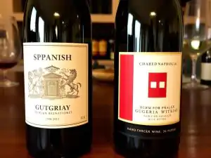 Dve tmavé fľaše vína na drevenom stole, ľavá fľaša má etiketu s nápisom "SPPANISH GUTGRIAY" a erbom, pravá fľaša má etiketu s červeným obdĺžnikom a nápisom "CHARED NAPHOLIA GUGERIA WITLIAS".